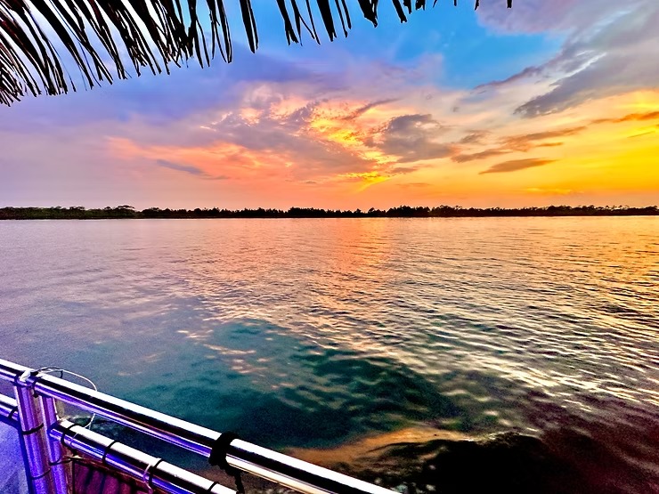 Colorful sunset over a calm lake with silhouetted trees and a thatched roof in the foreground.