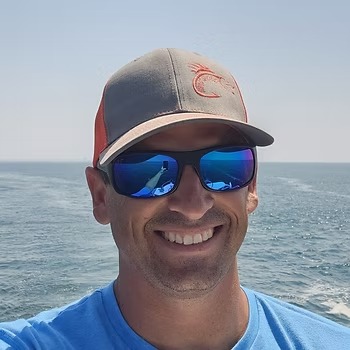 Man in sunglasses and cap smiling by the sea on a sunny day.