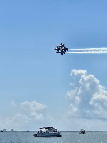 Four jets flying in formation above a bay with boats and blue sky.