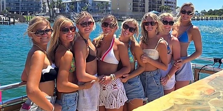 Eight women in swimsuits and sunglasses on a boat with water and buildings in the background.