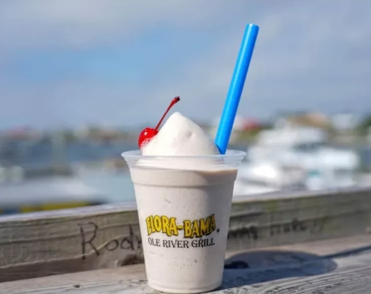 Frozen drink with a cherry and blue straw on a wooden surface, ocean view in background.