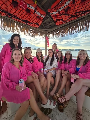 Group of women in pink shirts seated on a boat, with life jackets above and water in the background.