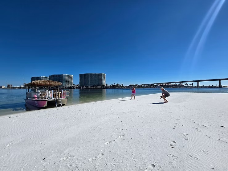 People playing on a sunny beach with a pontoon boat and buildings in the background.