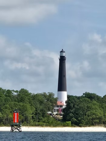 Tall black and white lighthouse with trees, beach, and small red tower in foreground.