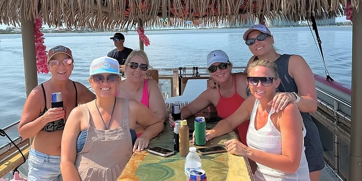 Six women relaxing at a tiki bar on a boat, with water in the background.
