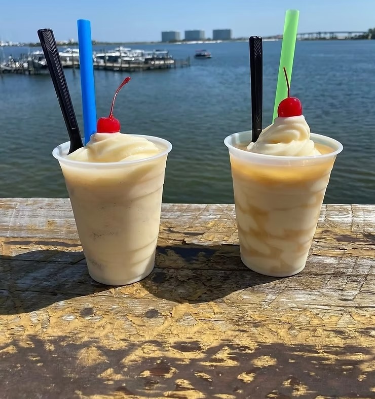 Two frozen drinks with cherries and colorful straws on a wooden table by a lake, under a clear blue sky.
