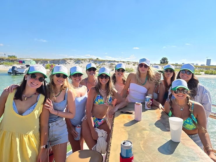 Group of women in swimsuits and hats smiling by water, holding drinks, with a sandy beach in the background.