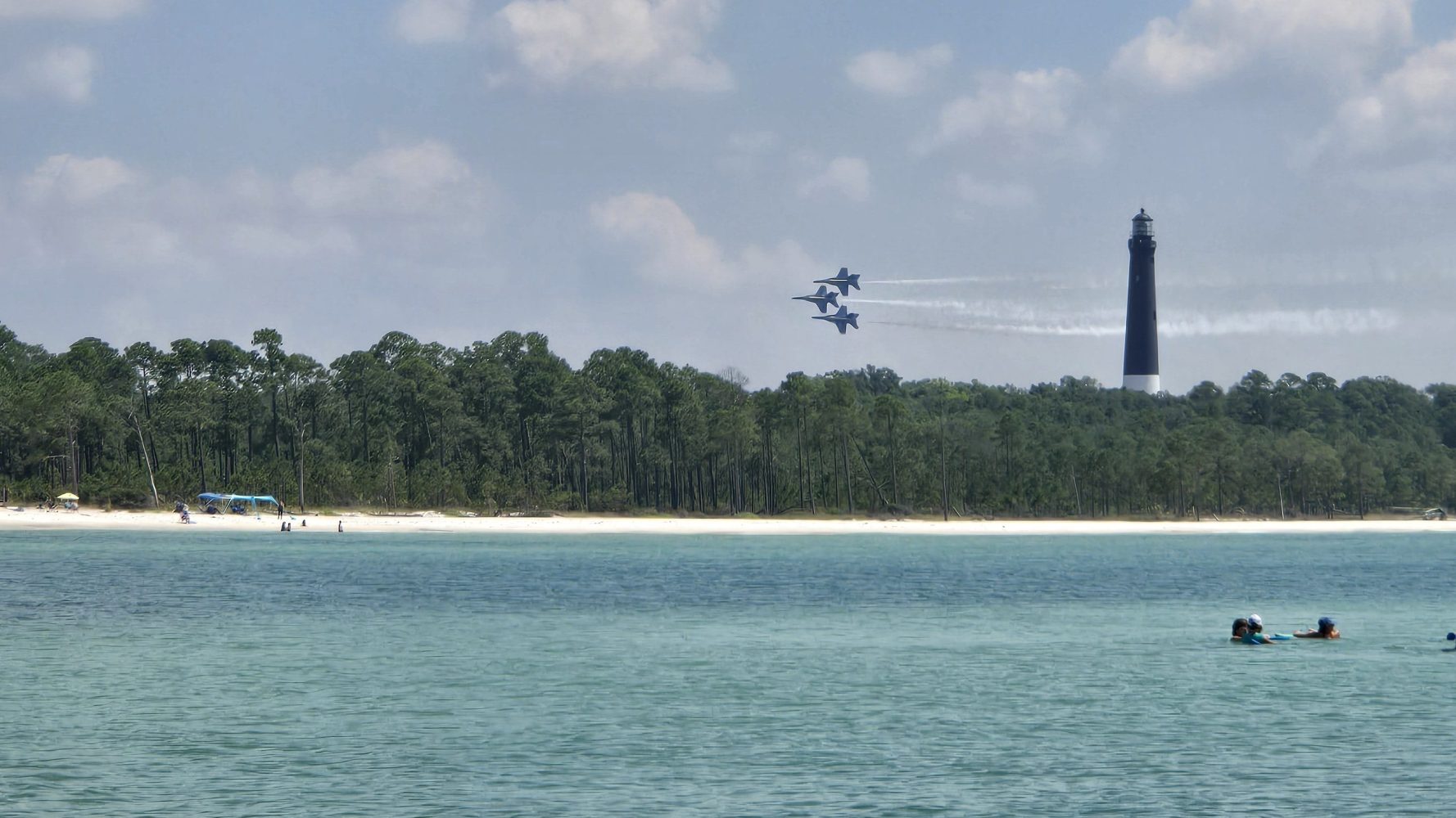 Blue Angel Jets flying near the tall Pensacola Lighthouse, above a beach with people and clear blue water.