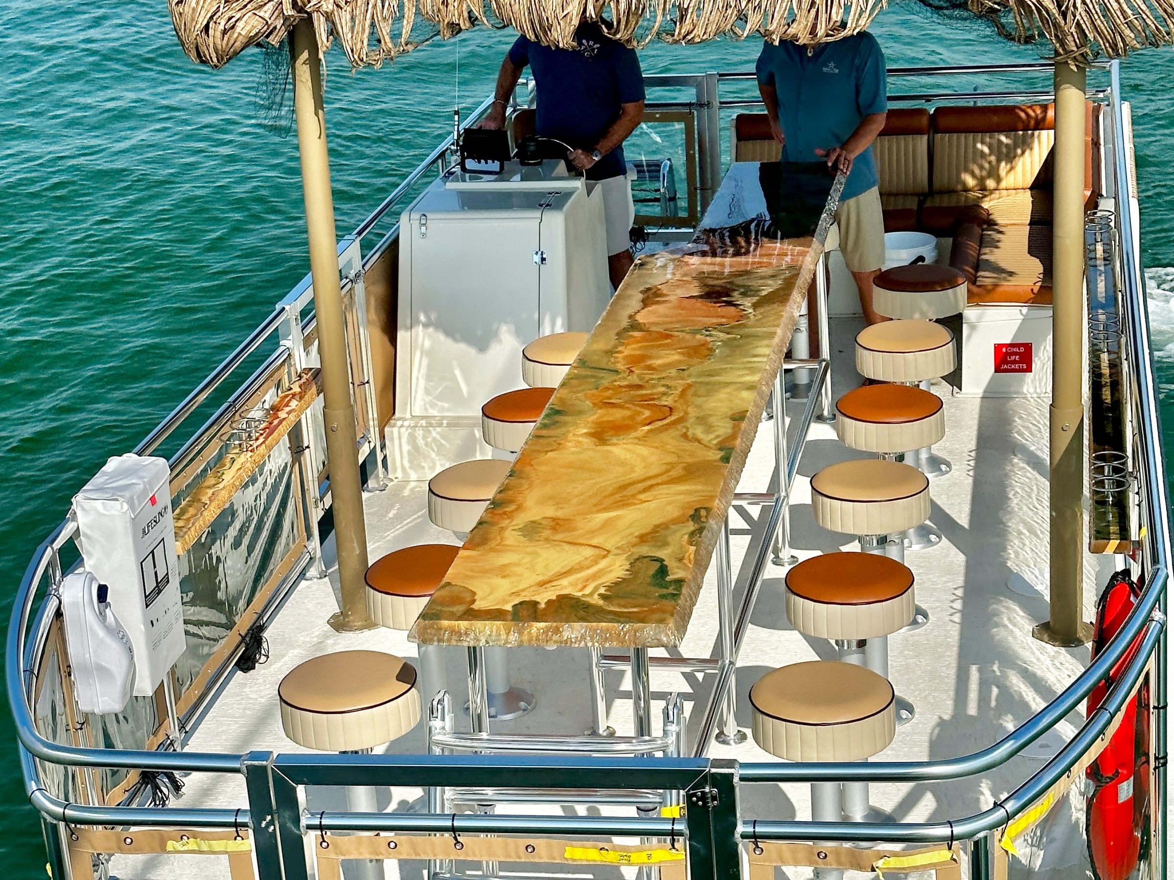 Boat with a tiki bar, thatched roof, stools, and people on the water.